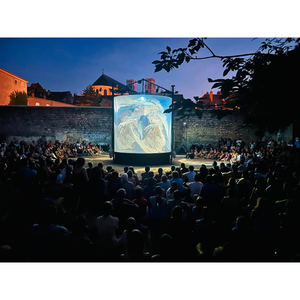 une foule ets assise en cerle, au centre un grand cylindre lumineux. Il fait nuit.
